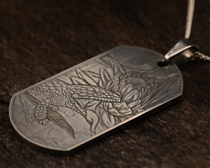 Hand‑engraved titanium pendant featuring a blue‑tongue lizard sunning beside a flower