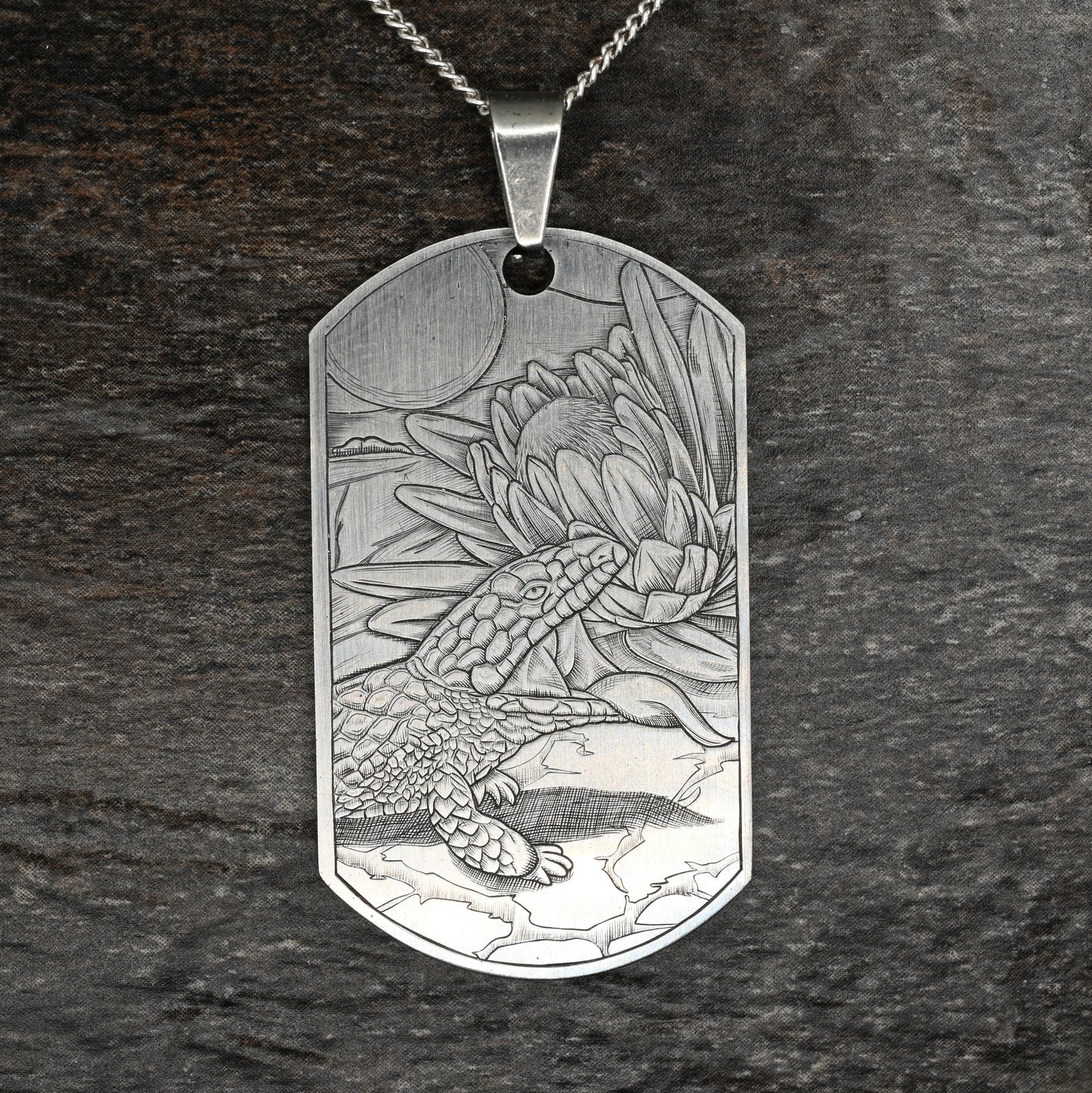 Hand‑engraved titanium pendant featuring a blue‑tongue lizard sunning beside a flower